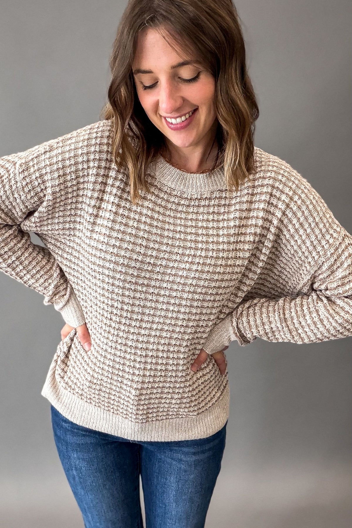 A woman wearing a beige and cream waffle-knit crewneck sweater with pockets, paired with blue jeans, posing against a solid gray studio background.
