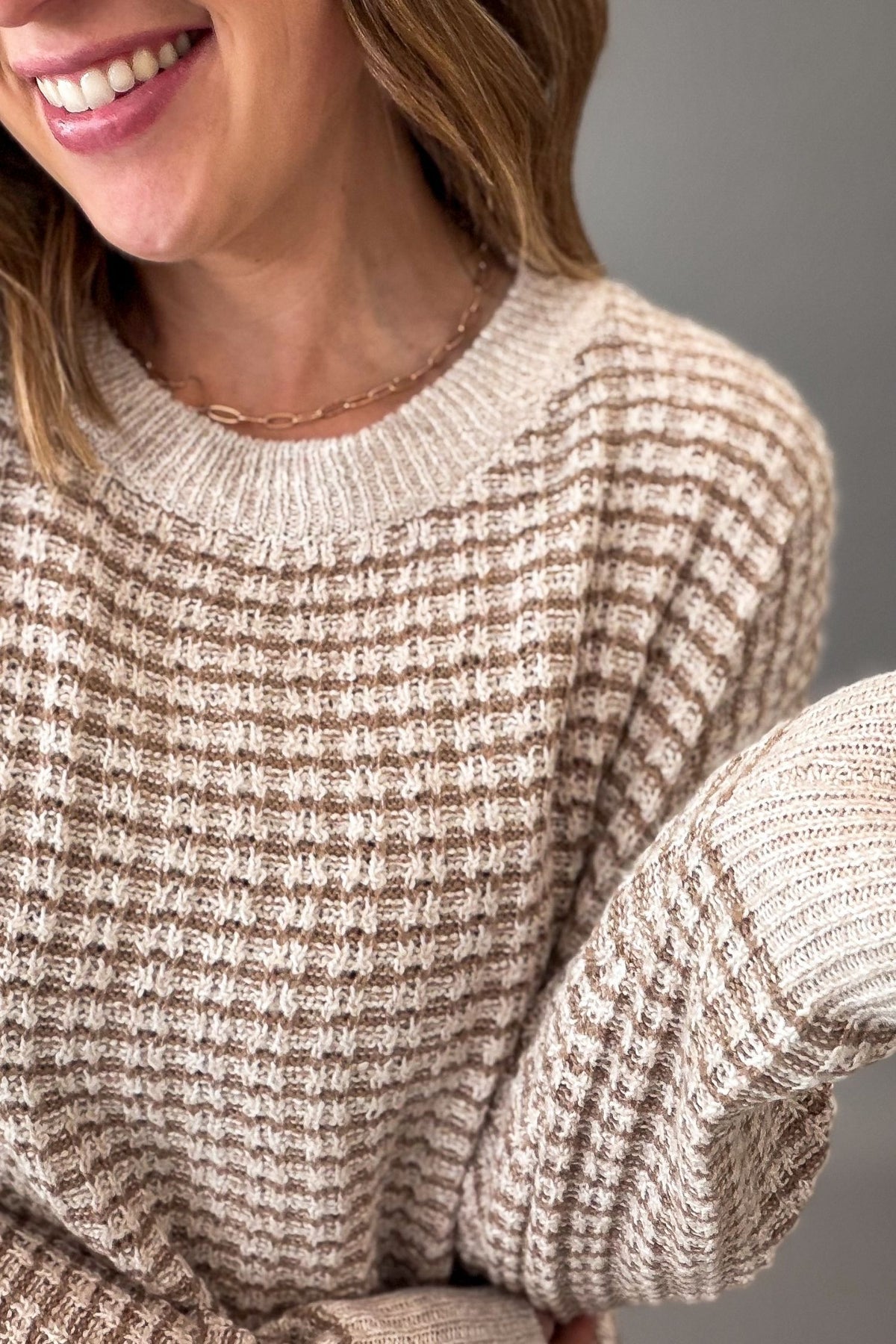A close-up of a woman wearing a chunky knit crewneck sweater with a textured waffle pattern in beige and brown.