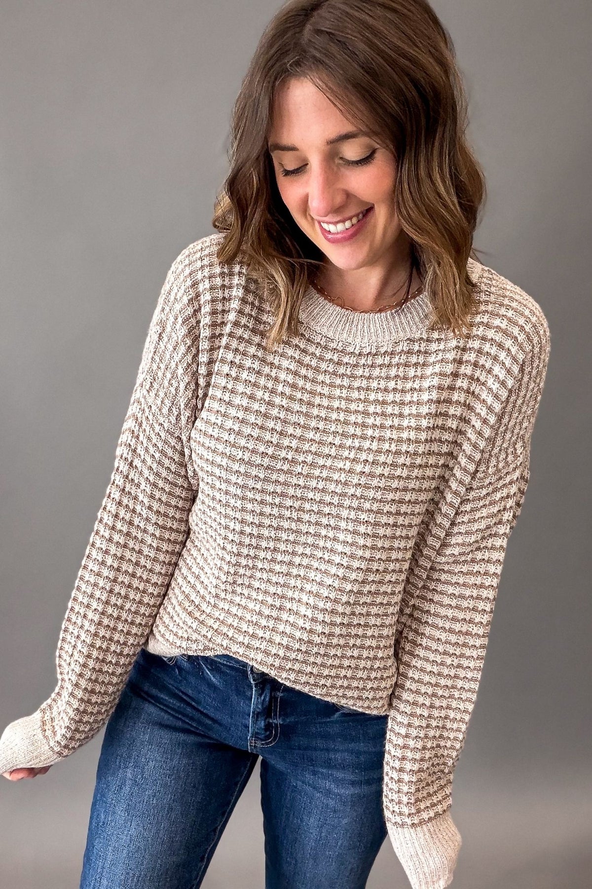 A woman wearing a beige and brown textured knit crewneck sweater with blue jeans, posing against a solid gray studio background.