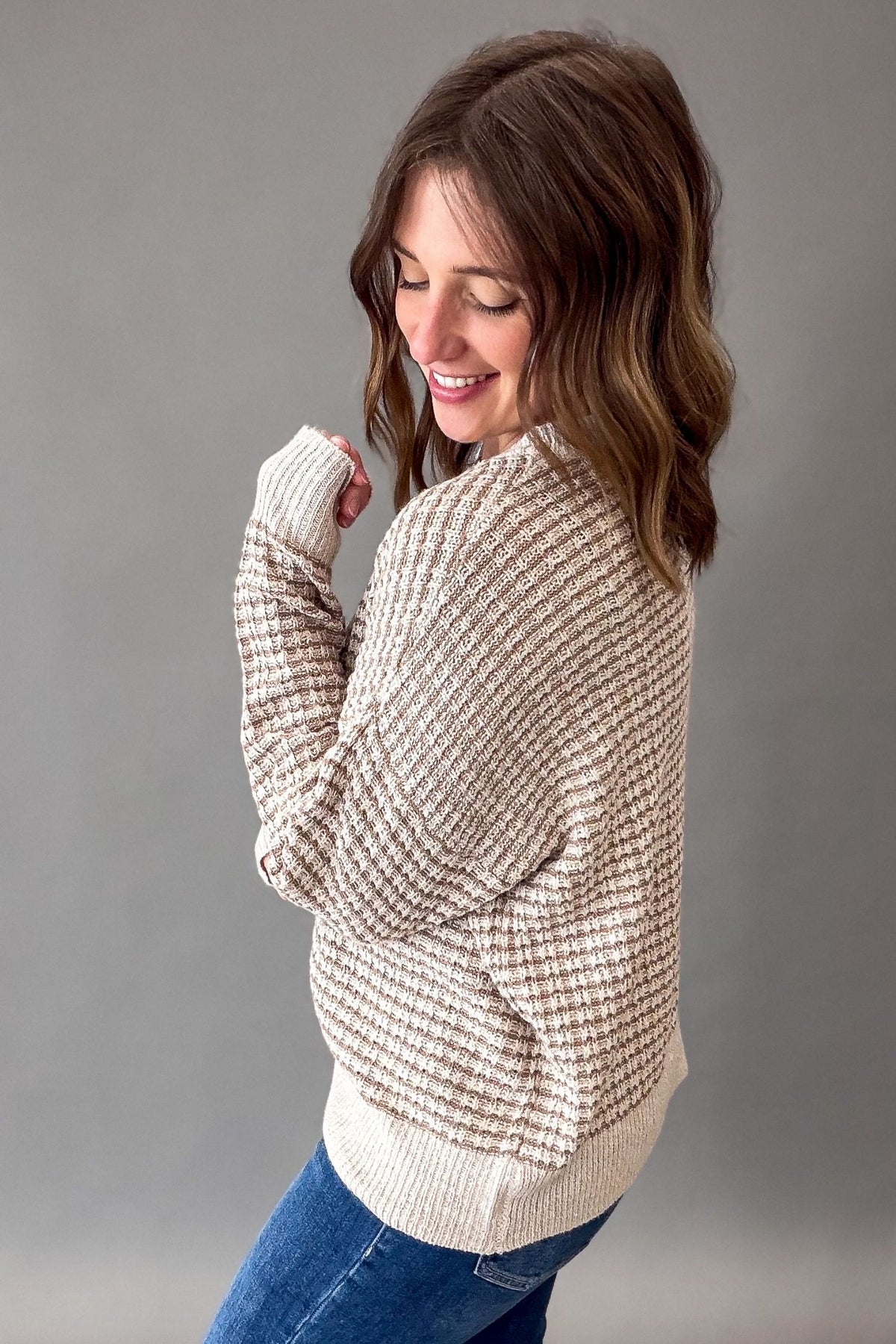 A woman wearing a textured cream and beige waffle-knit sweater with blue jeans, photographed against a solid gray studio background.