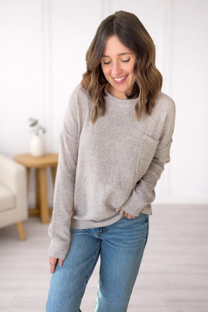 Woman wearing a taupe textured sweater and blue jeans in a room with a white wall and wooden table.