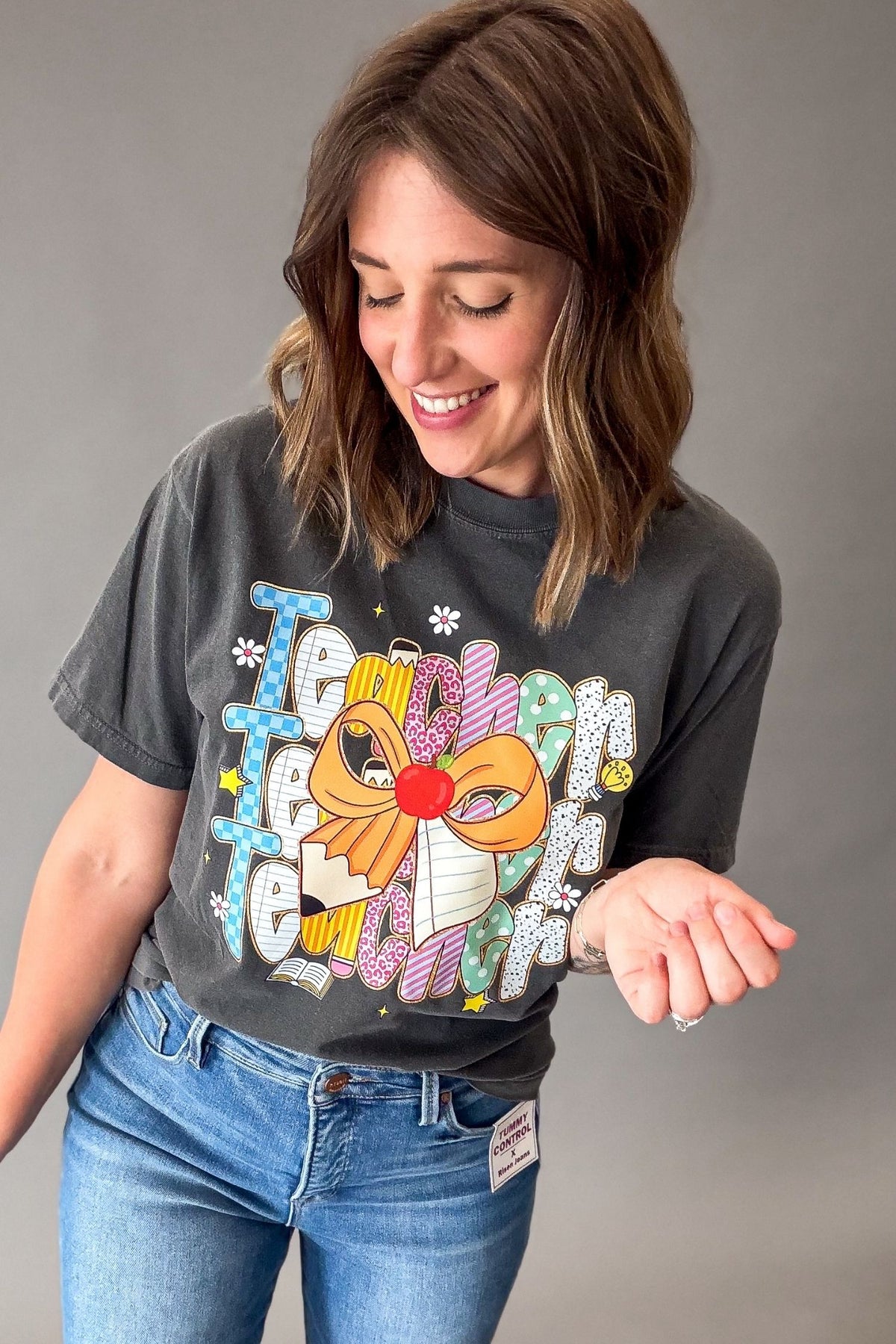 A woman wearing a dark charcoal gray graphic t-shirt with a colorful pencil bow and 'Teacher' design, paired with blue jeans, against a solid gray studio background.