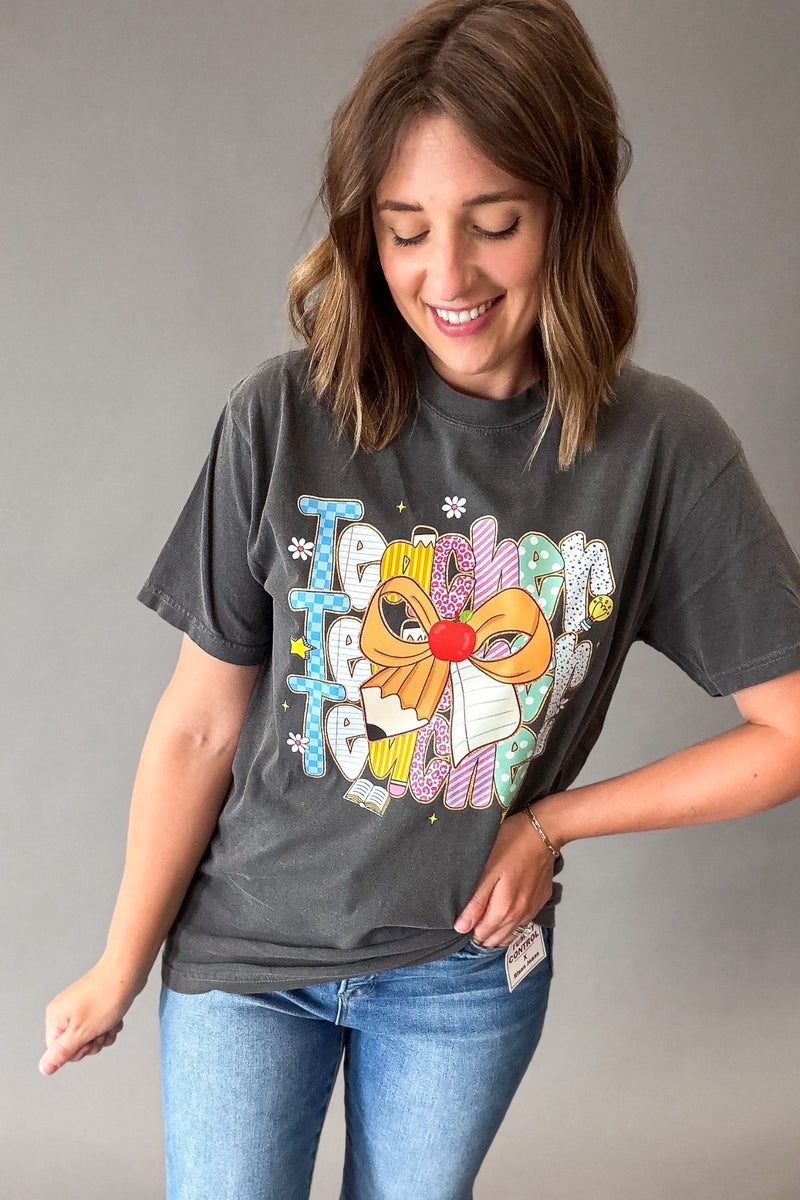 A woman wearing a dark gray graphic t-shirt with a colorful teacher graphic and pencil bow and the word 'Teacher' on the front, paired with blue jeans, standing against a plain gray background.