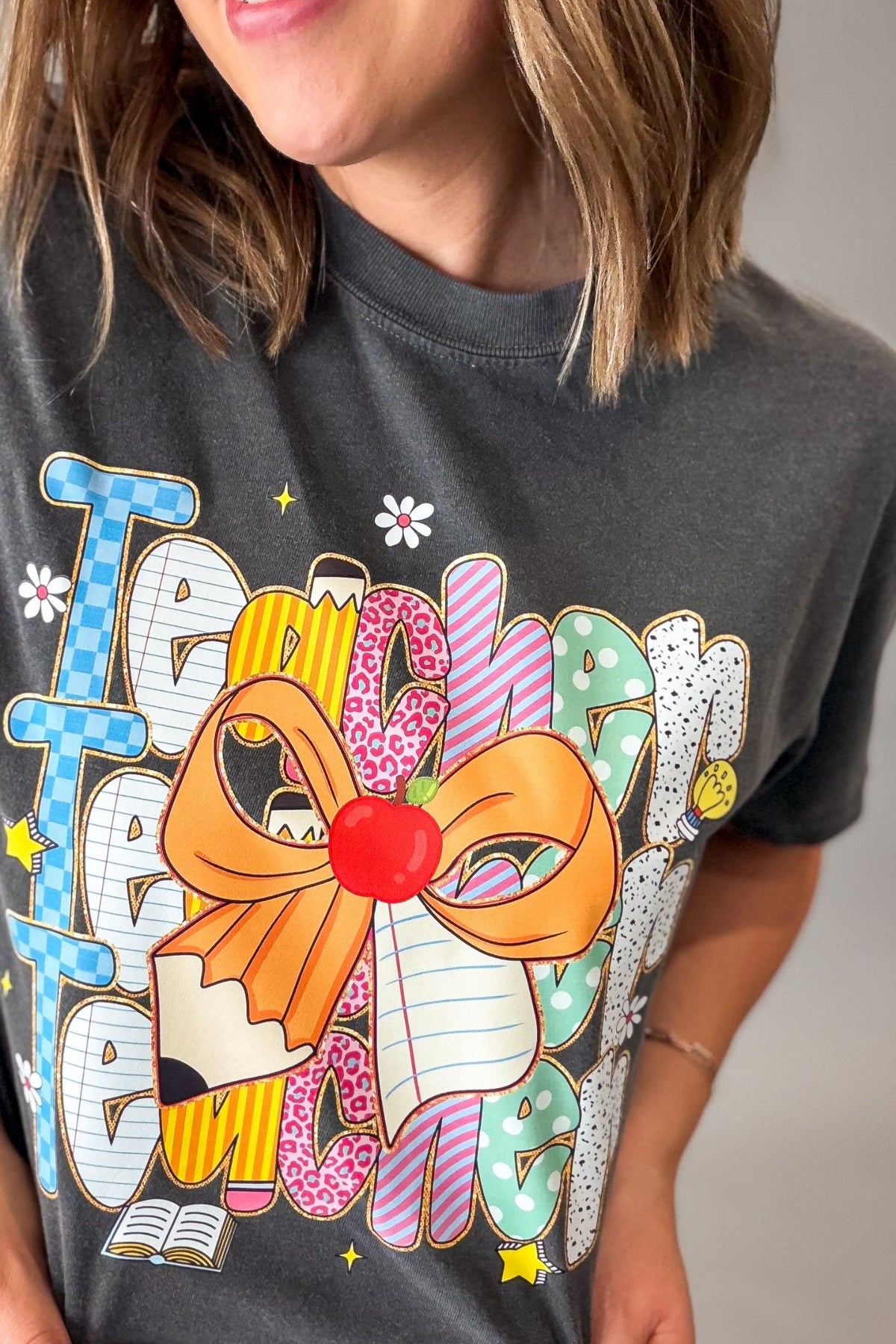 A close-up of a person wearing a dark gray graphic t-shirt with a colorful retro-style 'Teacher' design featuring a large orange bow, pencil, and patterned letters.