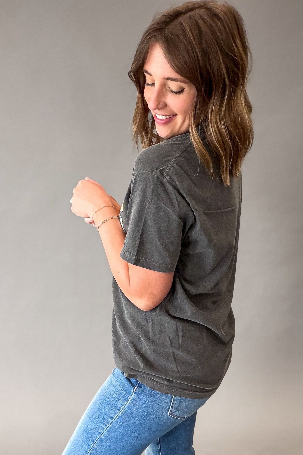 A woman wearing a dark charcoal gray short-sleeve t-shirt and blue jeans, shown from a side-back angle against a solid gray studio background.