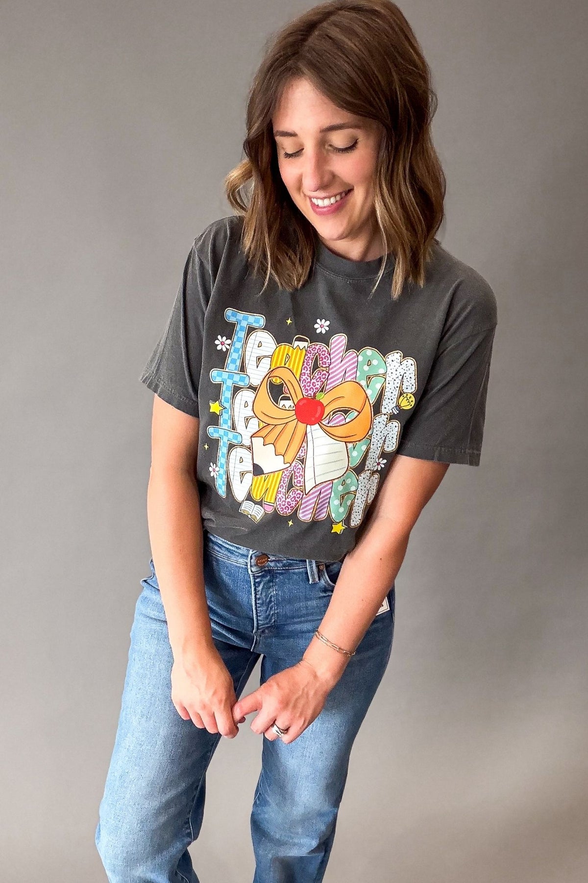 A woman wearing a dark gray graphic t-shirt with a colorful teacher design and blue jeans, standing against a plain gray studio background.