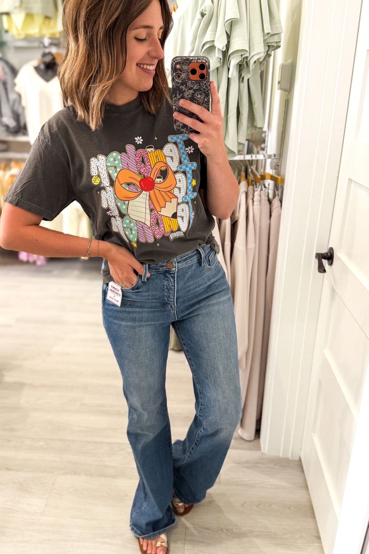 A woman takes a mirror selfie in a clothing store, wearing a dark charcoal graphic t-shirt with a colorful teacher design and blue jeans.