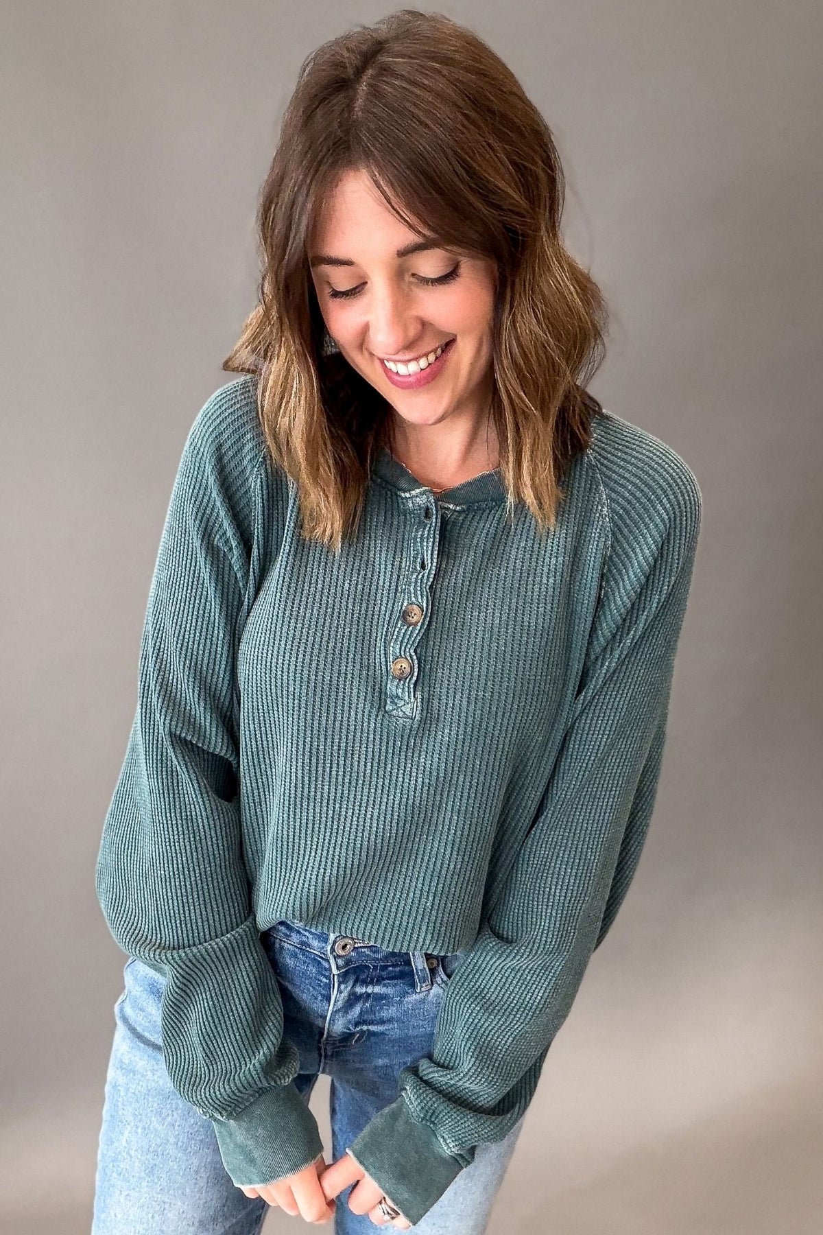 A woman wearing a sage green ribbed long-sleeve henley top and light wash blue jeans, smiling and looking down against a neutral gray studio background.