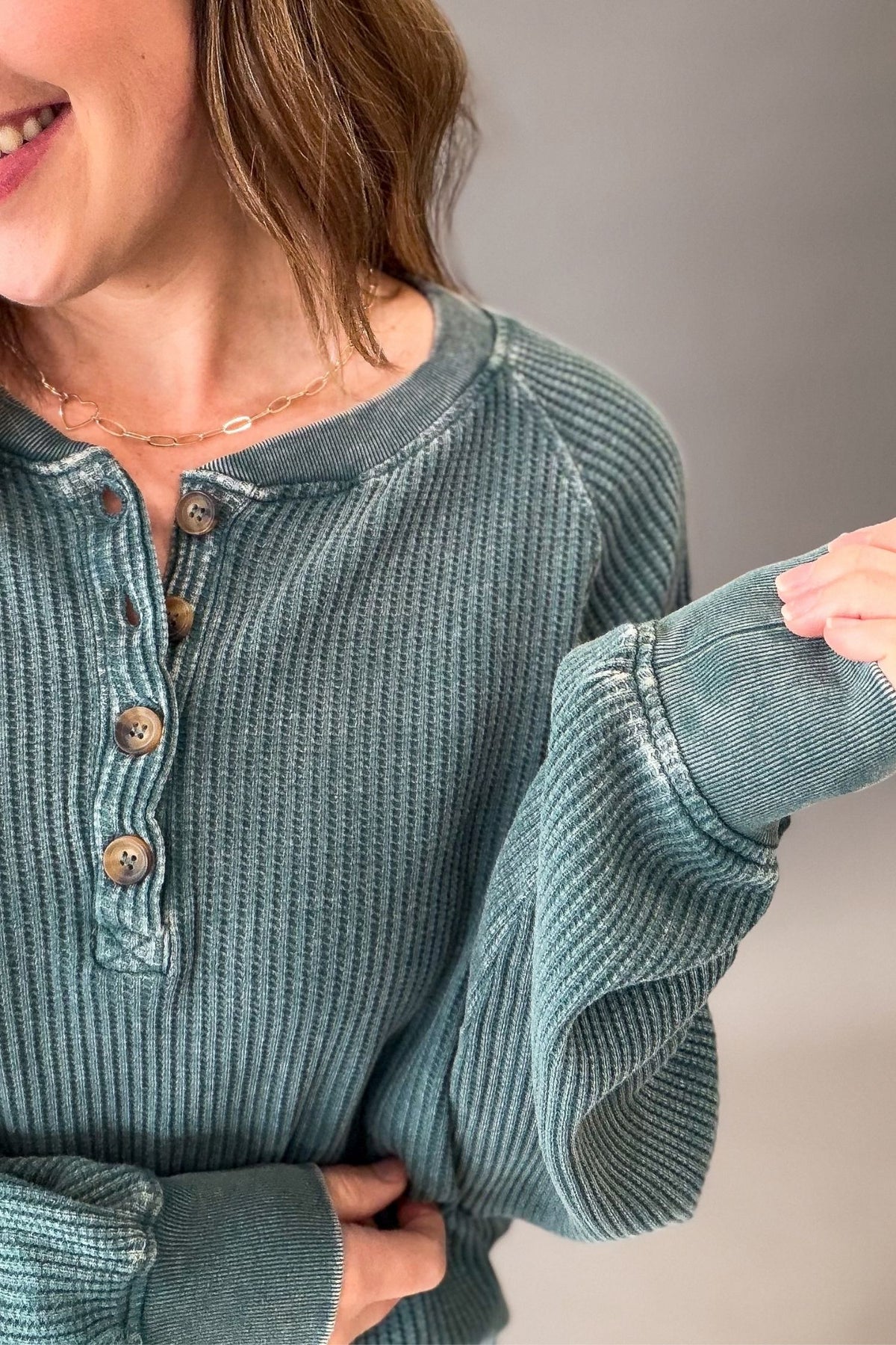 A close-up of a woman wearing a ribbed long-sleeve henley top in a muted teal-green color, showing the texture of the fabric and the brown buttons on the chest.