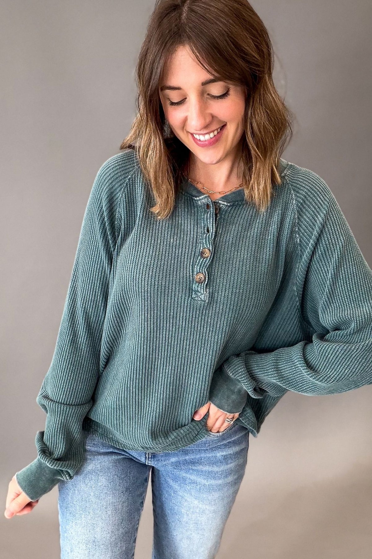 A woman wearing a ribbed long-sleeve henley top in muted teal blue with light wash jeans, posing against a neutral gray studio background.