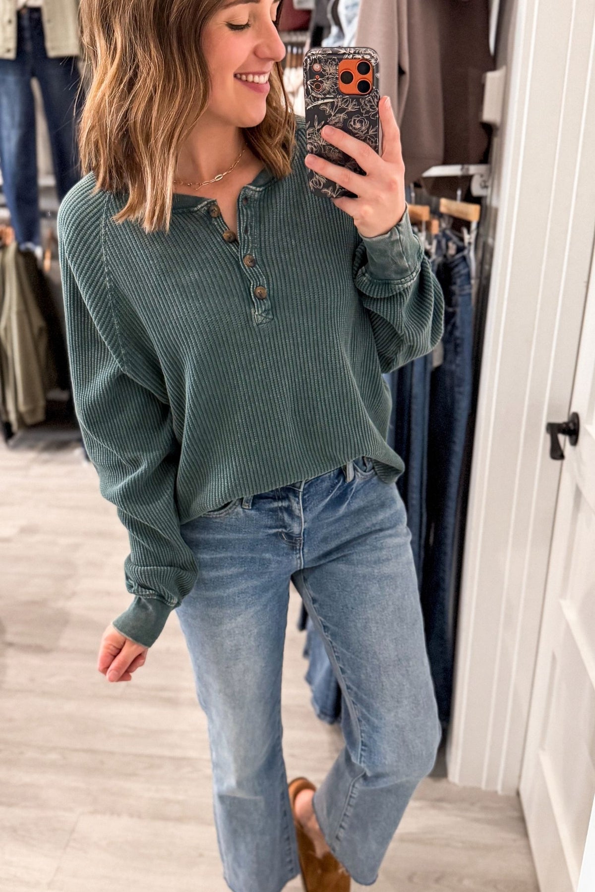 A woman takes a mirror selfie in a clothing store, wearing a sage green ribbed long-sleeve henley top and light-wash blue jeans.