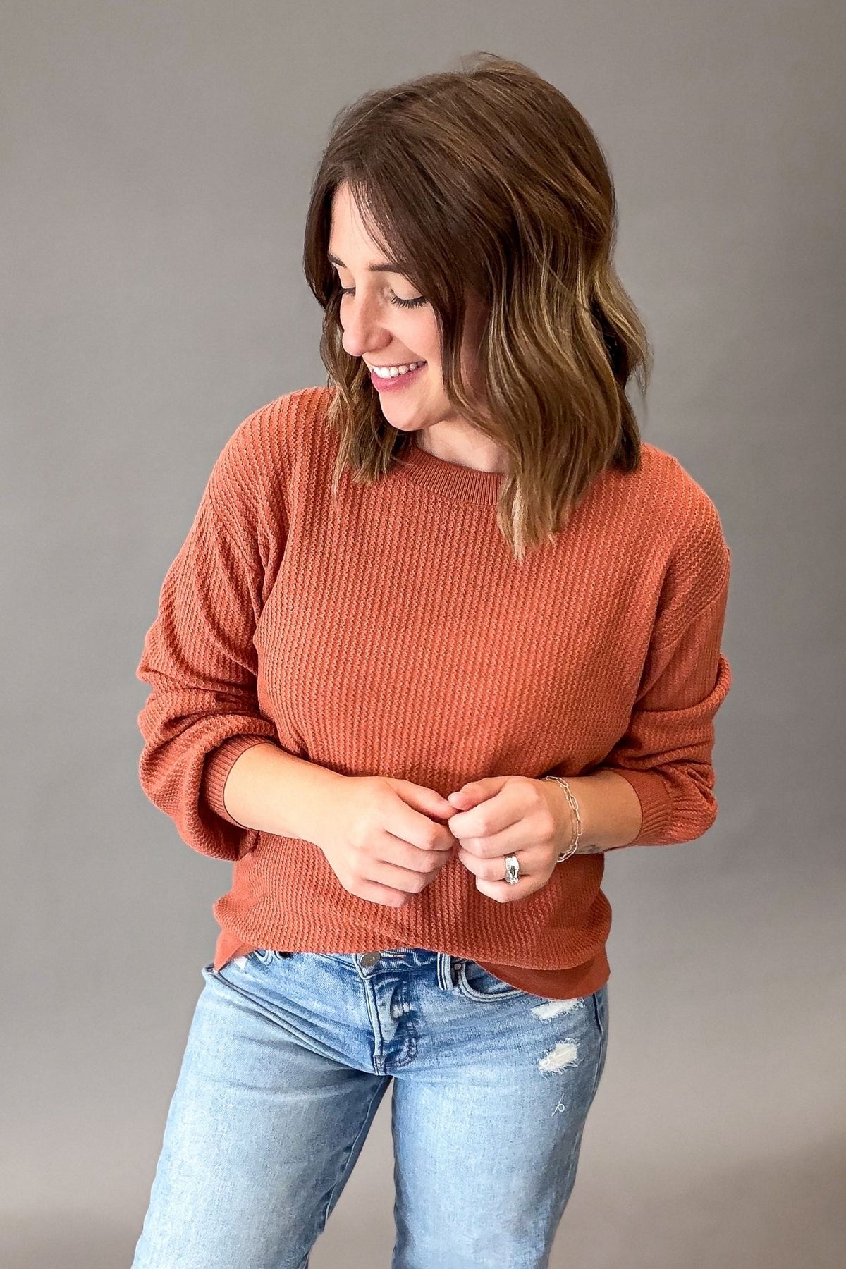 A woman wearing a rust-colored ribbed knit long-sleeve sweater and light blue jeans, standing against a solid gray studio background.