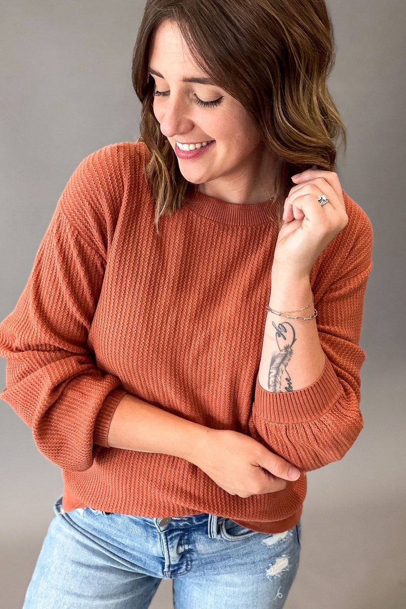 A woman wearing a rust-orange ribbed knit crewneck sweater and light-wash blue jeans, posing against a neutral gray studio background.