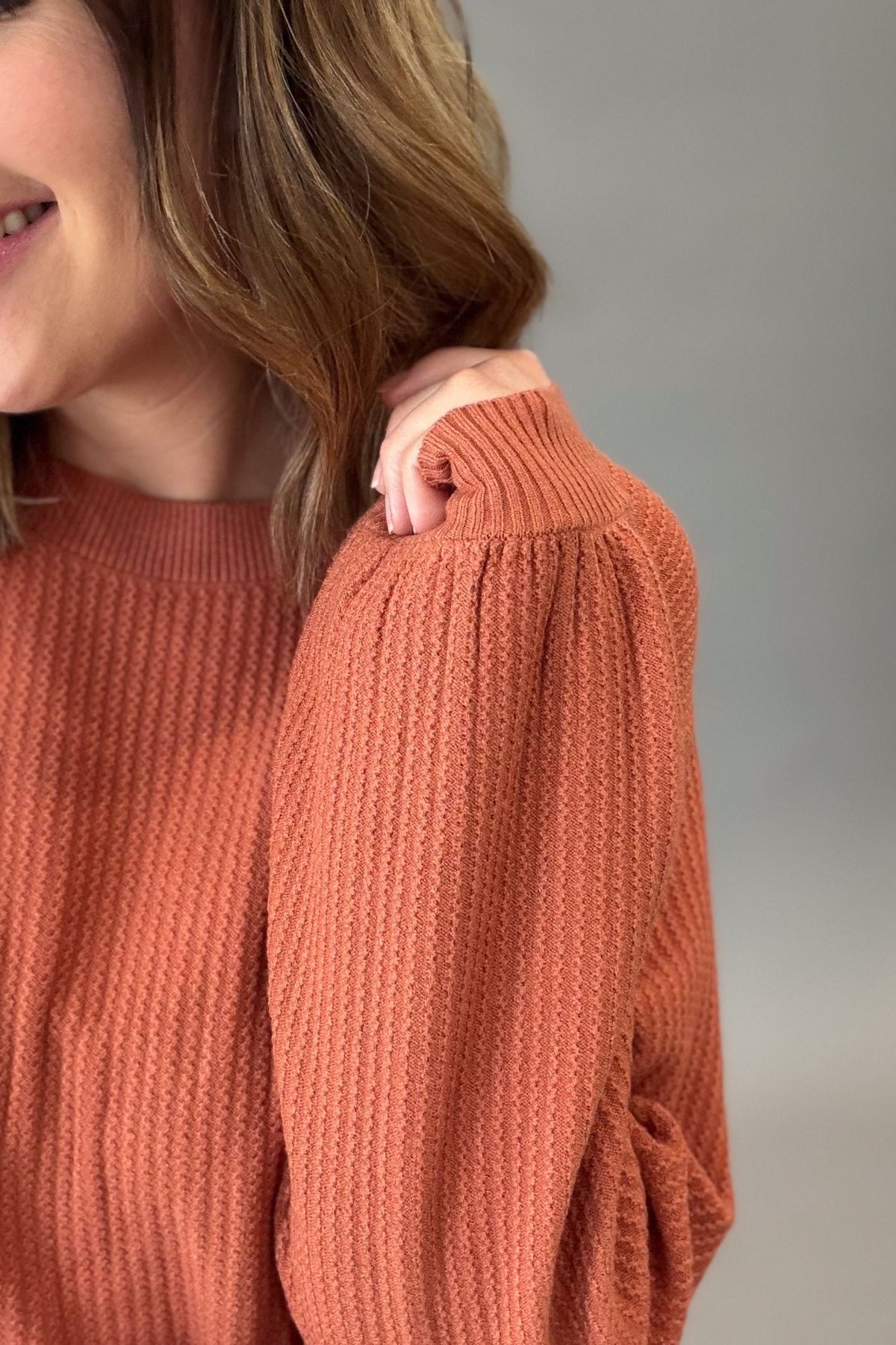 Close-up of a woman wearing a textured rust knit sweater, showing the ribbed crew neck and voluminous long sleeves against a plain gray background.