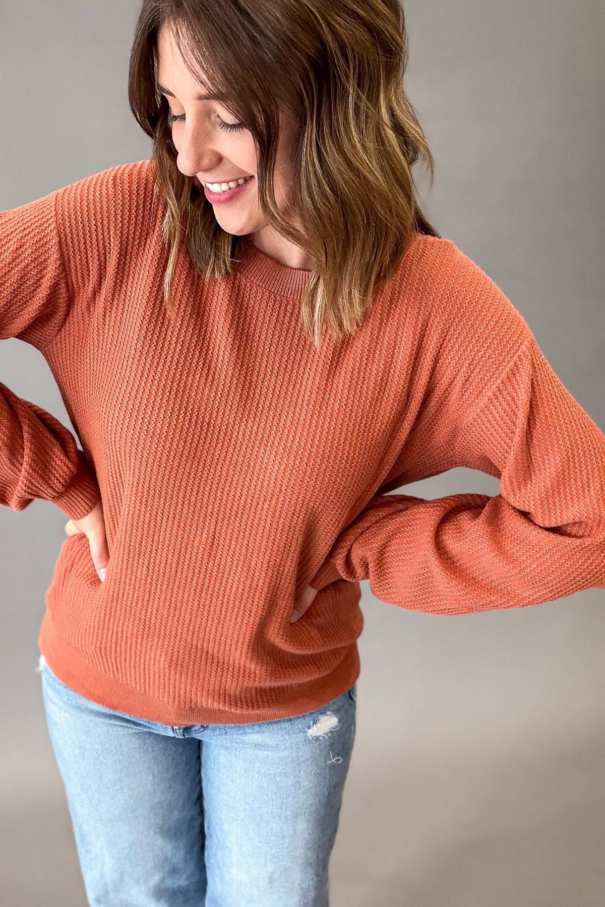 A woman wearing a rust-orange ribbed knit long-sleeve sweater and light-wash blue jeans, posing with her hands on her hips against a neutral gray studio background.