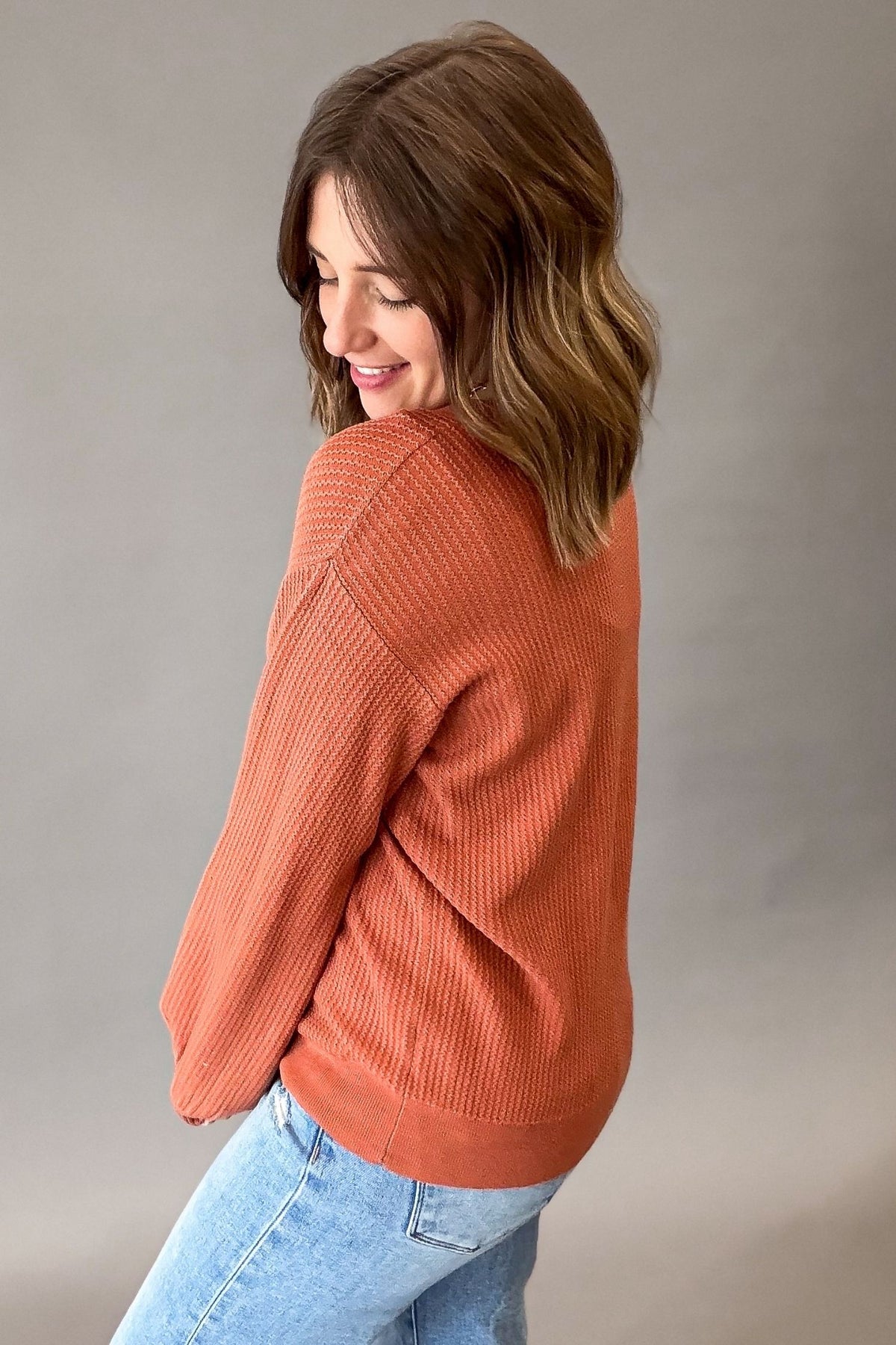 A woman wearing a ribbed knit long-sleeve sweater in a rust color, paired with light blue jeans, photographed against a neutral gray studio background.