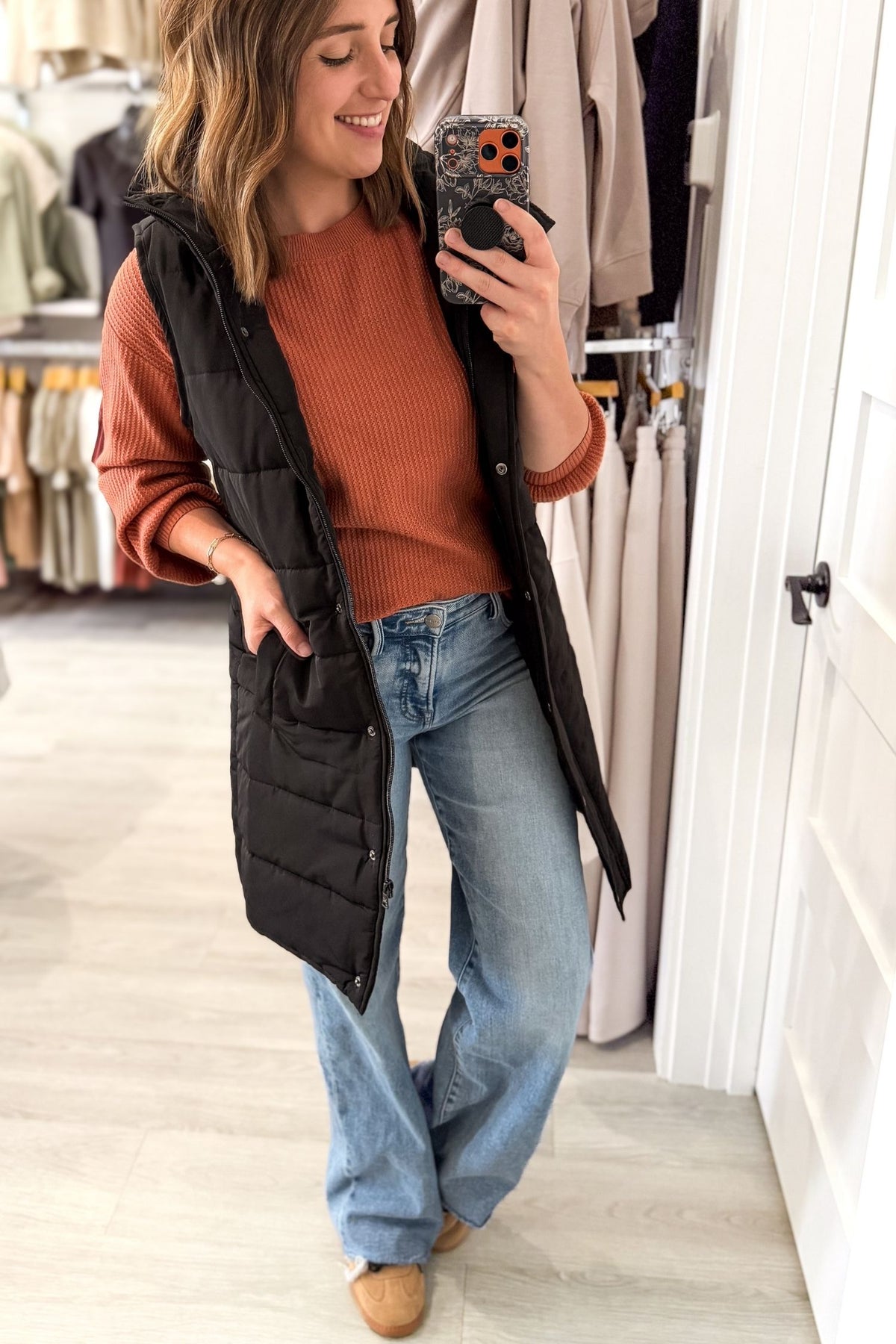 A woman takes a mirror selfie in a clothing boutique, wearing a black quilted puffer vest over a rust-orange ribbed sweater, blue wide-leg jeans, and tan slip-on shoes.