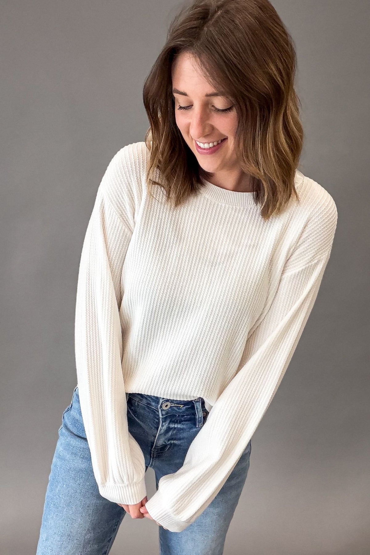 A woman wearing a white ribbed long-sleeve knit sweater and blue jeans, standing against a solid gray studio background.