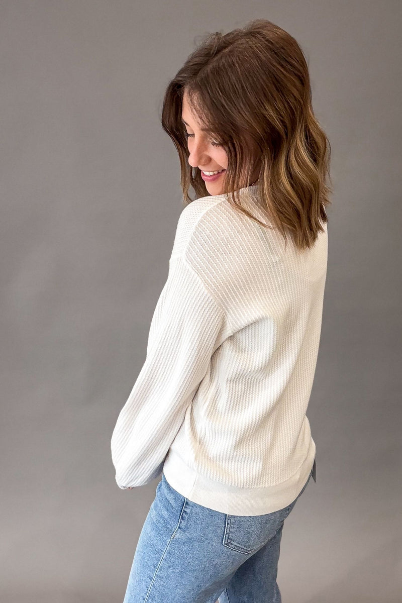 A woman with shoulder-length brown hair is shown from the back wearing a white ribbed knit sweater and light blue jeans against a solid gray studio background.