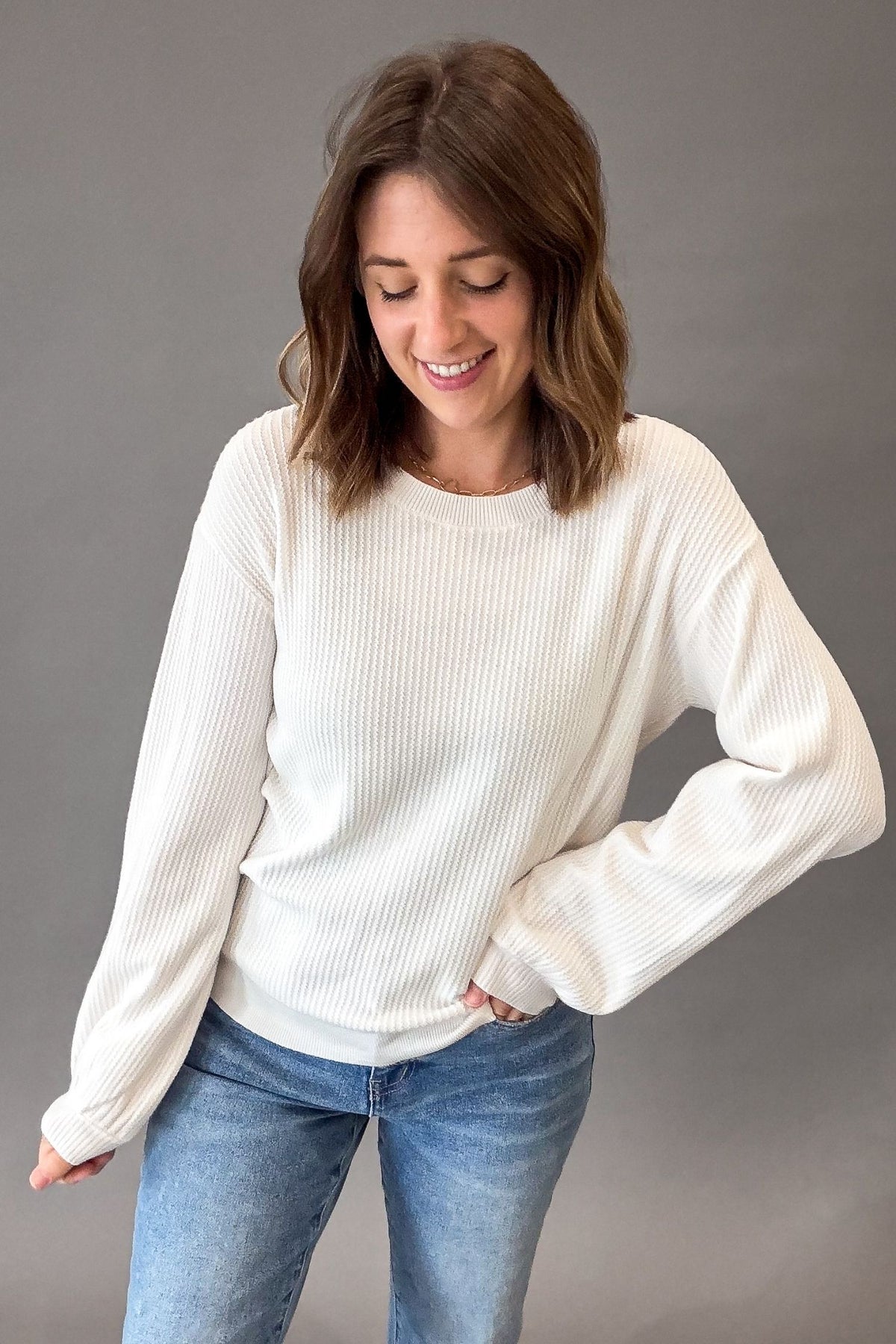 A woman wearing a white ribbed long-sleeve crewneck sweater and blue jeans, standing against a solid gray studio background.