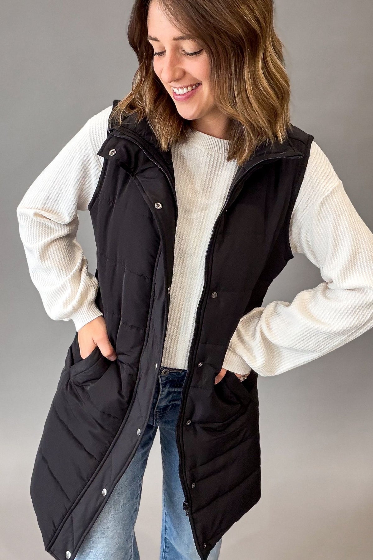 A woman wearing a black sleeveless puffer vest over a white ribbed sweater and blue jeans, standing against a plain gray studio background.