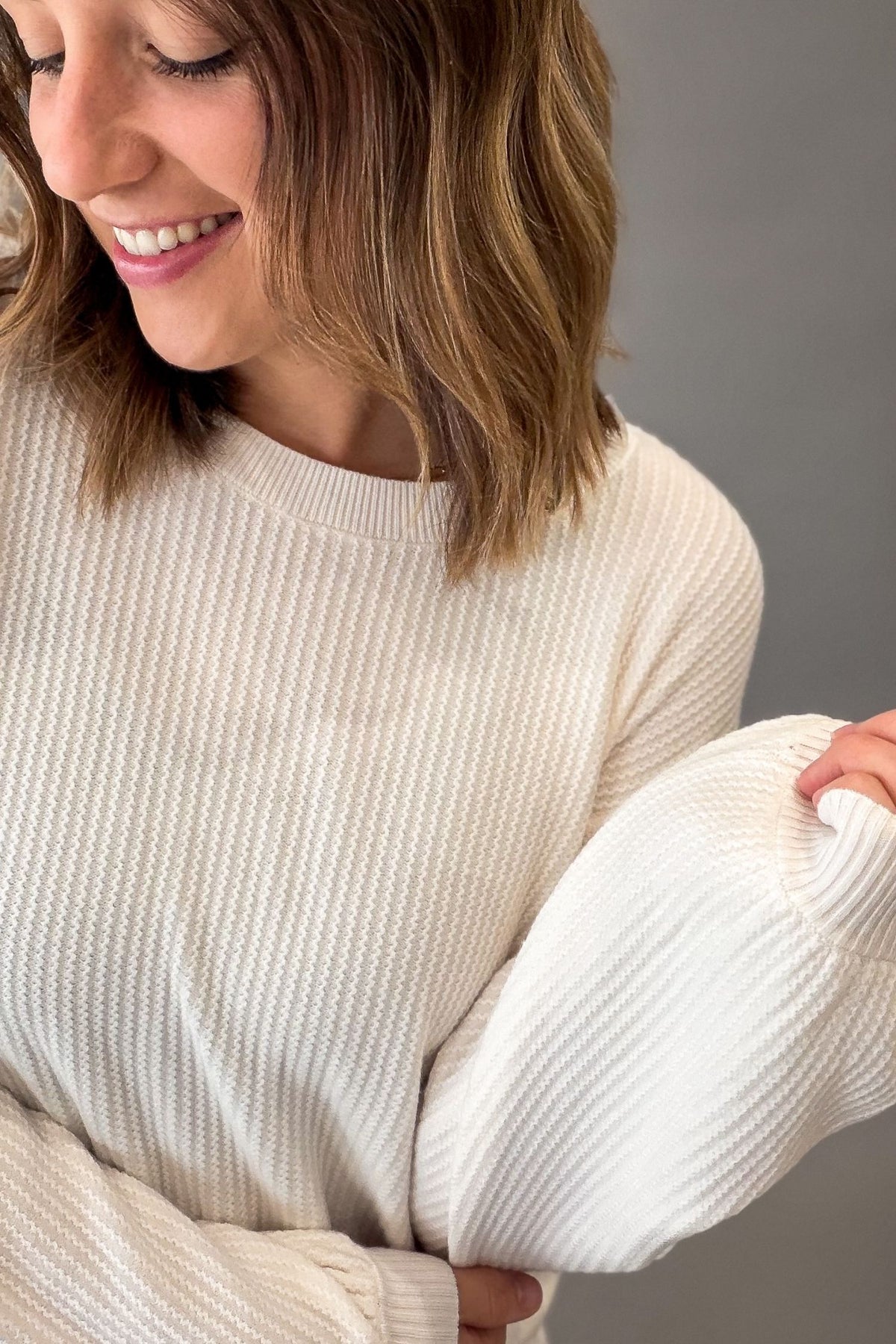 A woman wearing a white ribbed knit sweater with voluminous balloon sleeves, shown in a close-up shot against a neutral gray background.