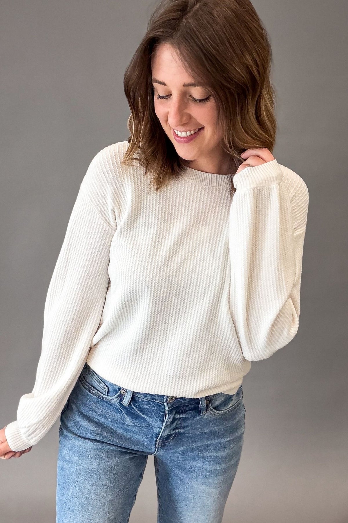 A woman wearing a white ribbed knit sweater with balloon sleeves and blue jeans, standing against a solid gray studio background.