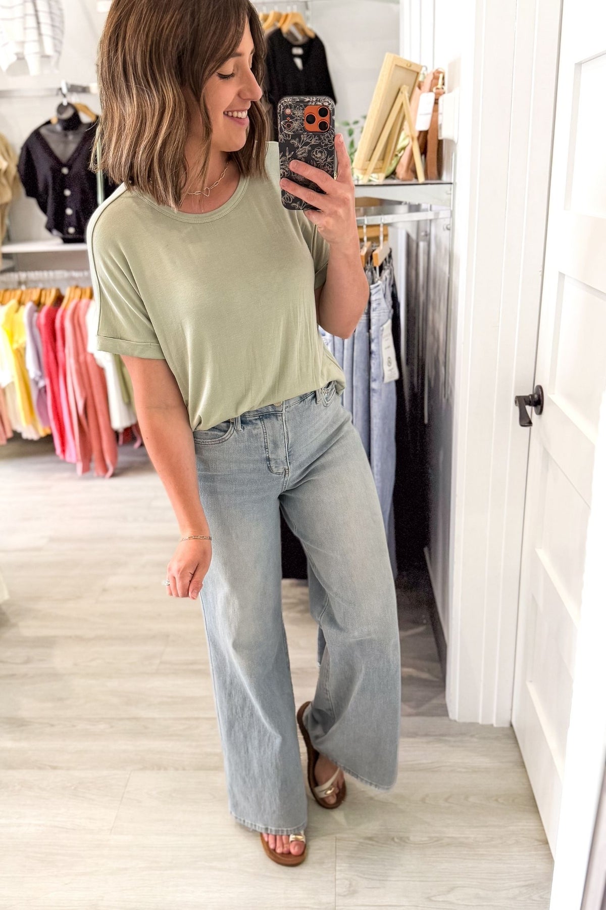 Woman taking a mirror selfie wearing a green t-shirt and light blue jeans in a clothing store.