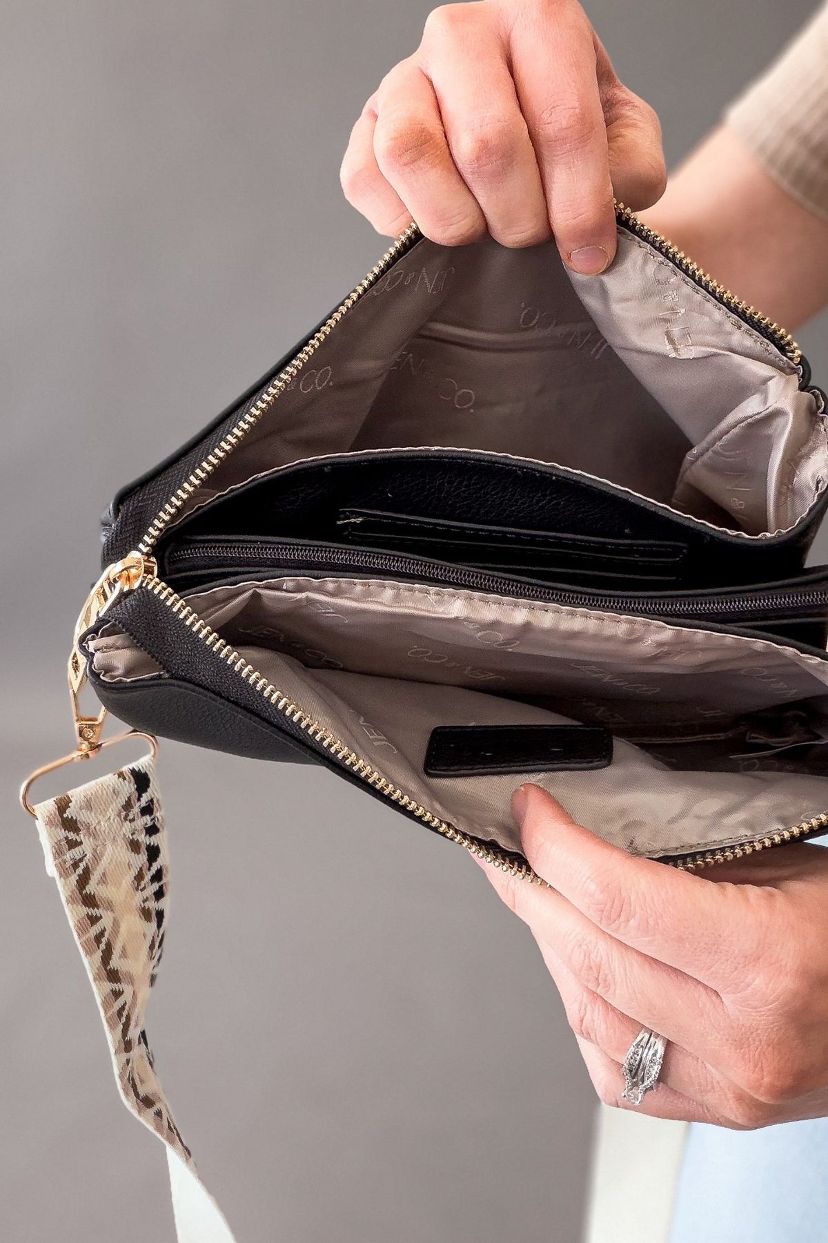 Person holding a black handbag with a patterned strap against a neutral background showing 2 larger compartments, one zipper compartment and card holders