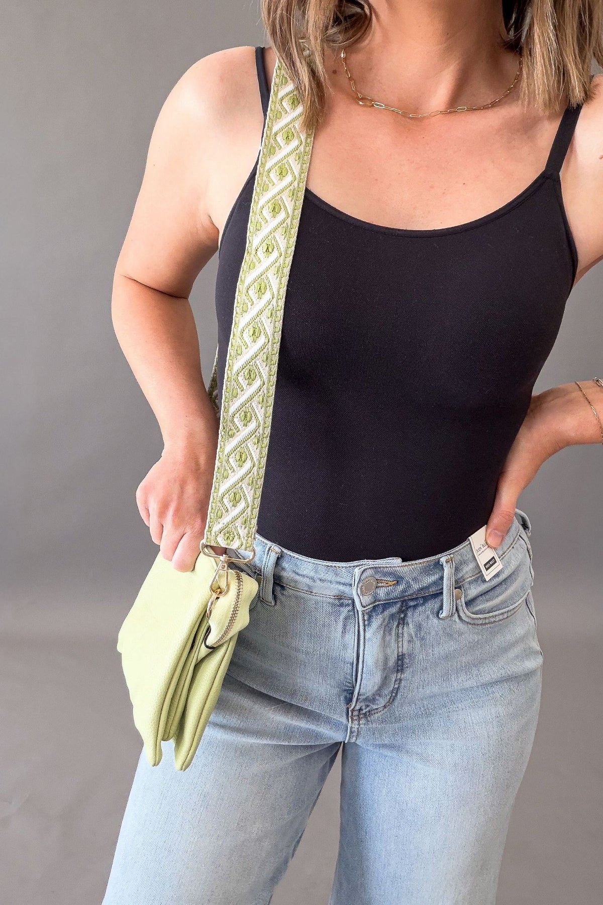 Person wearing a black tank top and light blue jeans, holding a green handbag with a patterned strap.