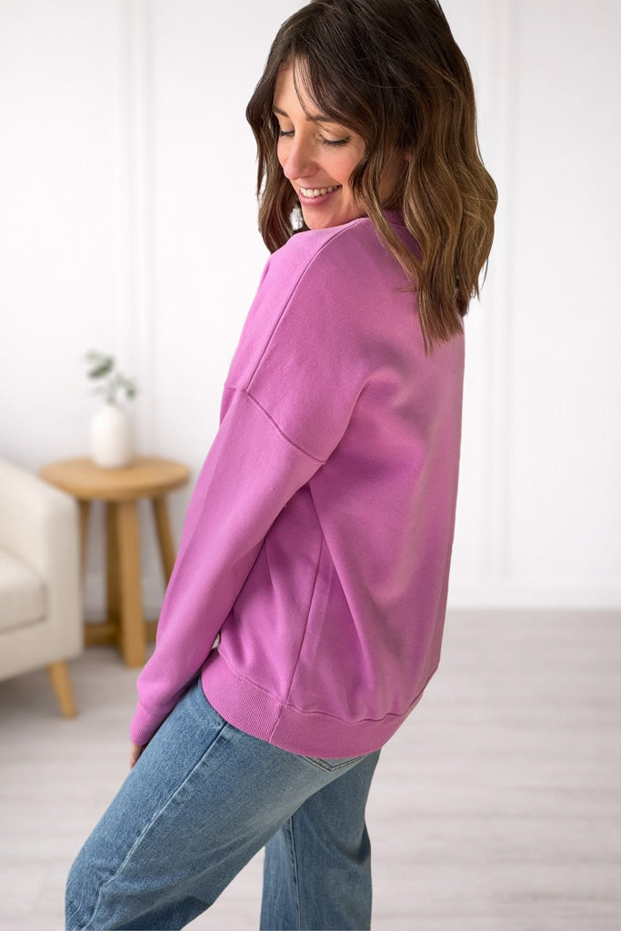 Woman wearing a mauve sweatshirt and blue jeans in a room with a white wall and wooden table.