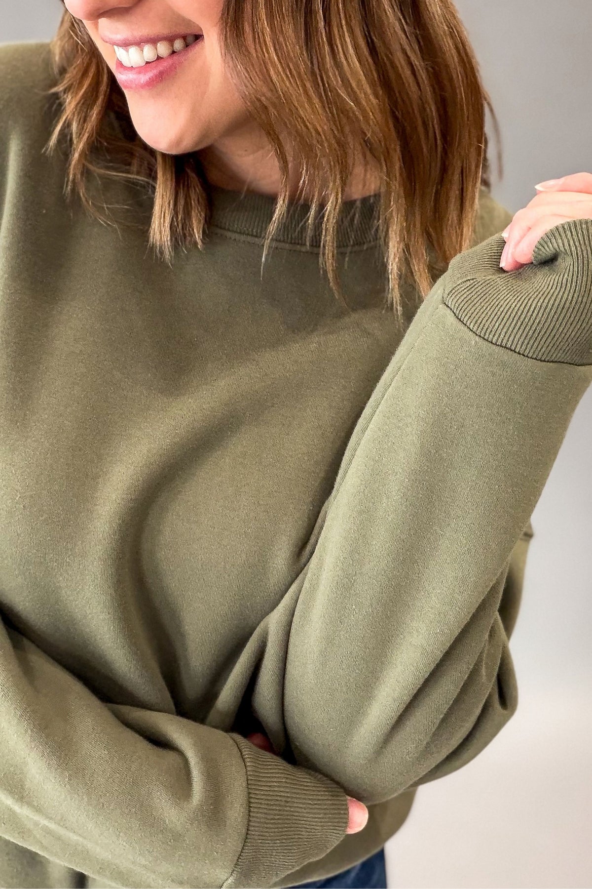 A close-up of a woman wearing an olive green crewneck sweatshirt, showing the ribbed cuffs and a portion of her smiling face.