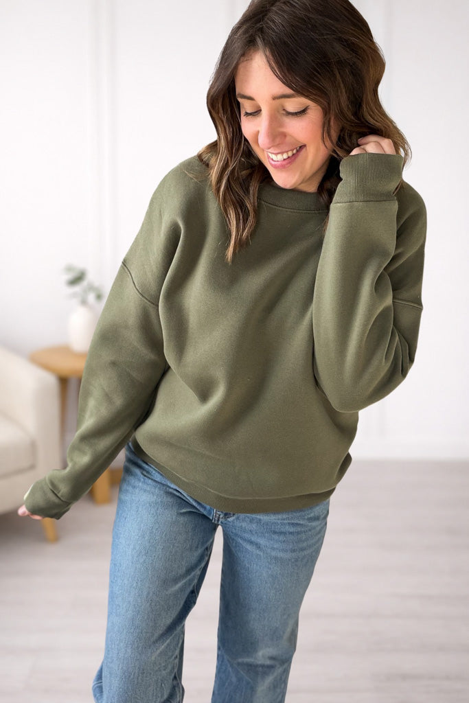 Woman wearing an olive green sweatshirt and blue jeans indoors.