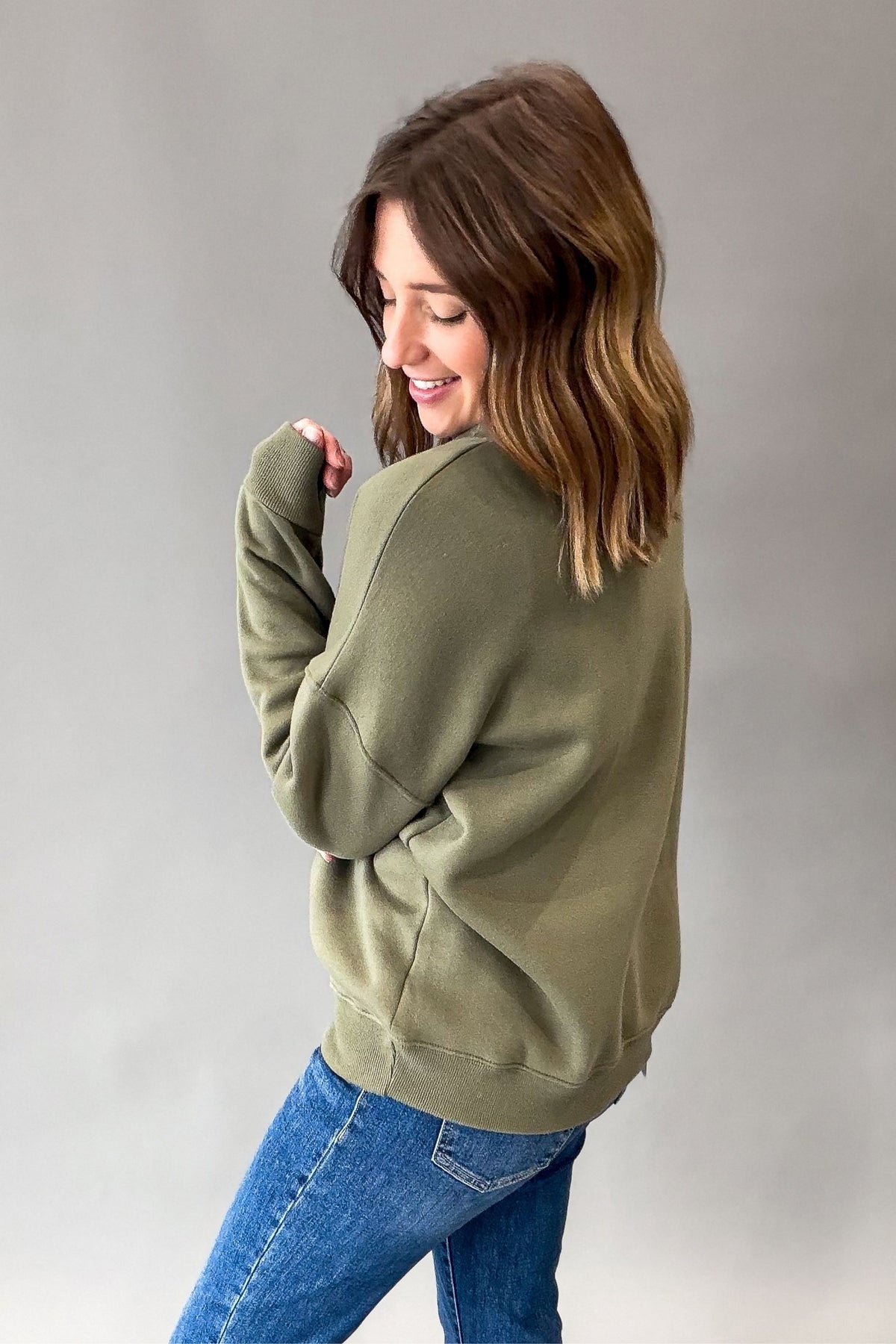 A woman wearing an olive green crewneck sweatshirt and blue jeans, shown from a side-back angle against a neutral gray studio background.