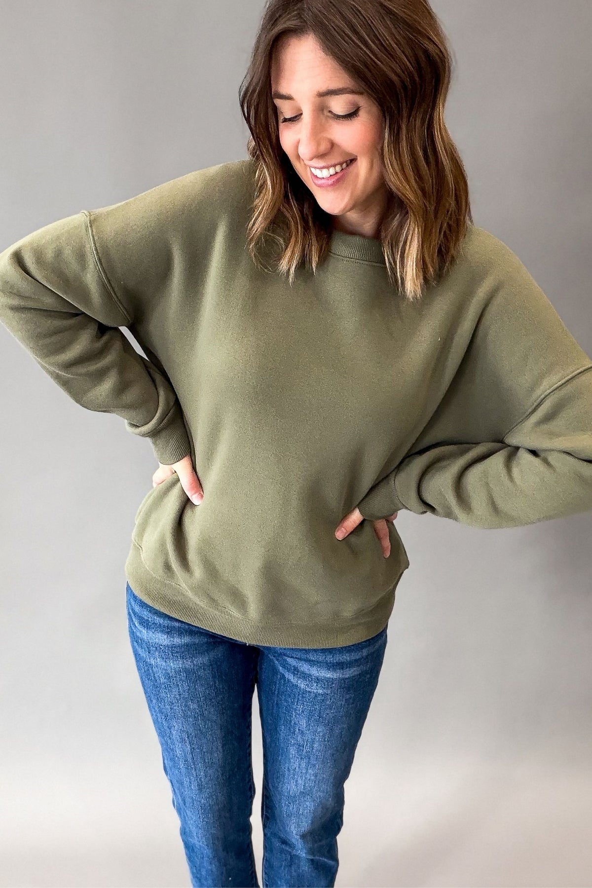 A woman wearing a relaxed olive green crewneck sweatshirt and blue jeans, standing against a neutral gray studio background.