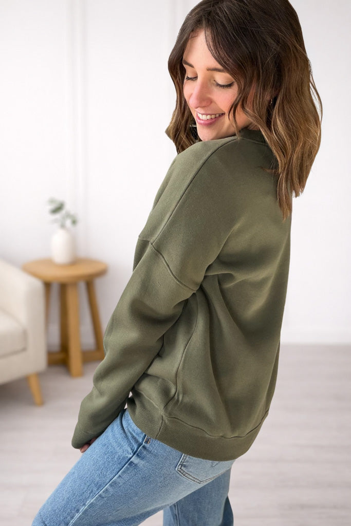 Woman wearing an olive green sweatshirt and blue jeans in a room with a white wall and wooden side table.