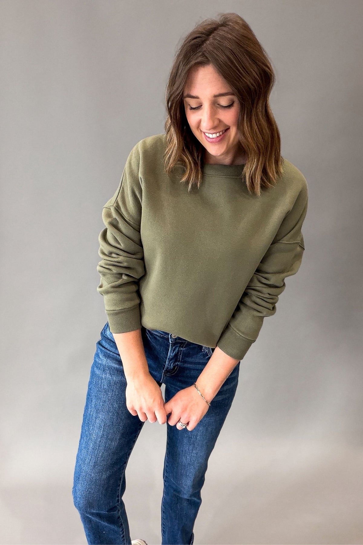 A woman wearing an olive green crewneck sweatshirt and blue jeans, standing against a neutral gray studio background.