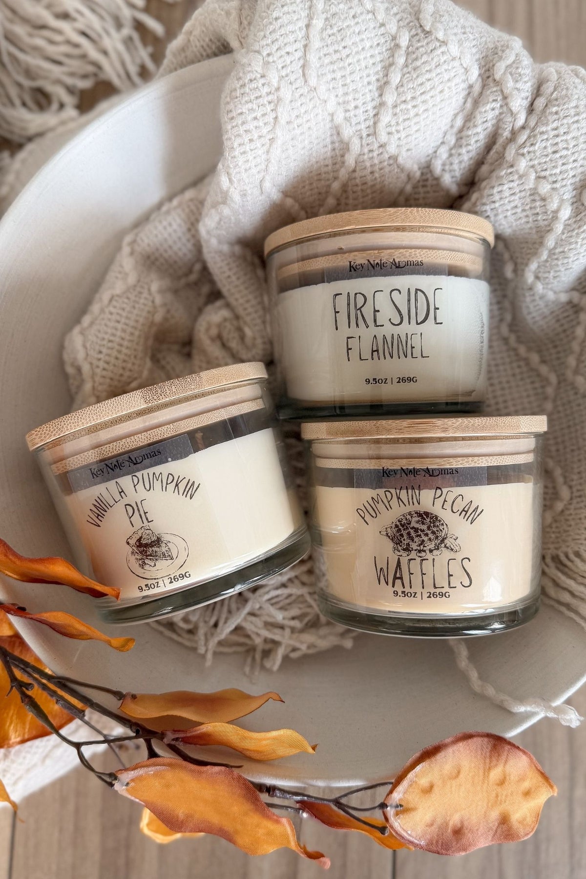 Three scented candles in glass jars with wooden lids are arranged on a white plate with a cream cable-knit blanket and fall leaves. The labels read Vanilla Pumpkin Pie, Fireside Flannel, and Pumpkin Pecan Waffles.