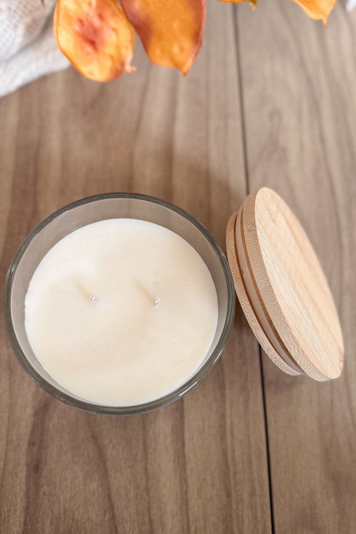 A two-wick soy candle in a clear glass jar with a light natural wood lid resting beside it on a wooden tabletop. The candle wax is a warm off-white/cream color.