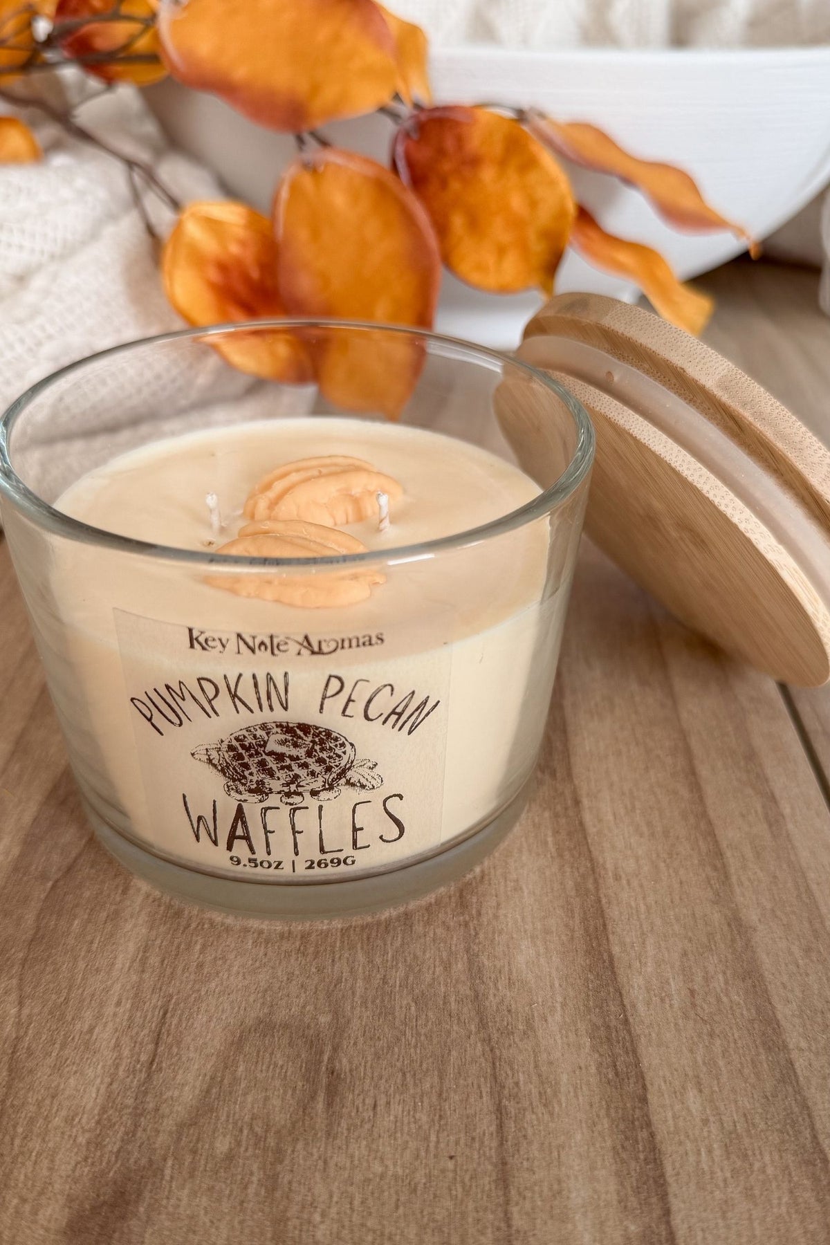 A glass jar candle with a light wood lid sits on a wooden surface. The candle wax is cream-colored with a printed label reading 'Key Note Aromas' and 'Pumpkin Pecan Waffles.
