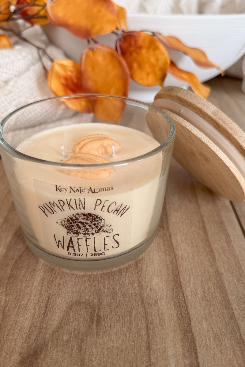 A glass jar candle with a light wood lid sits on a wooden surface. The candle wax is cream-colored with a printed label reading 'Key Note Aromas' and 'Pumpkin Pecan Waffles.