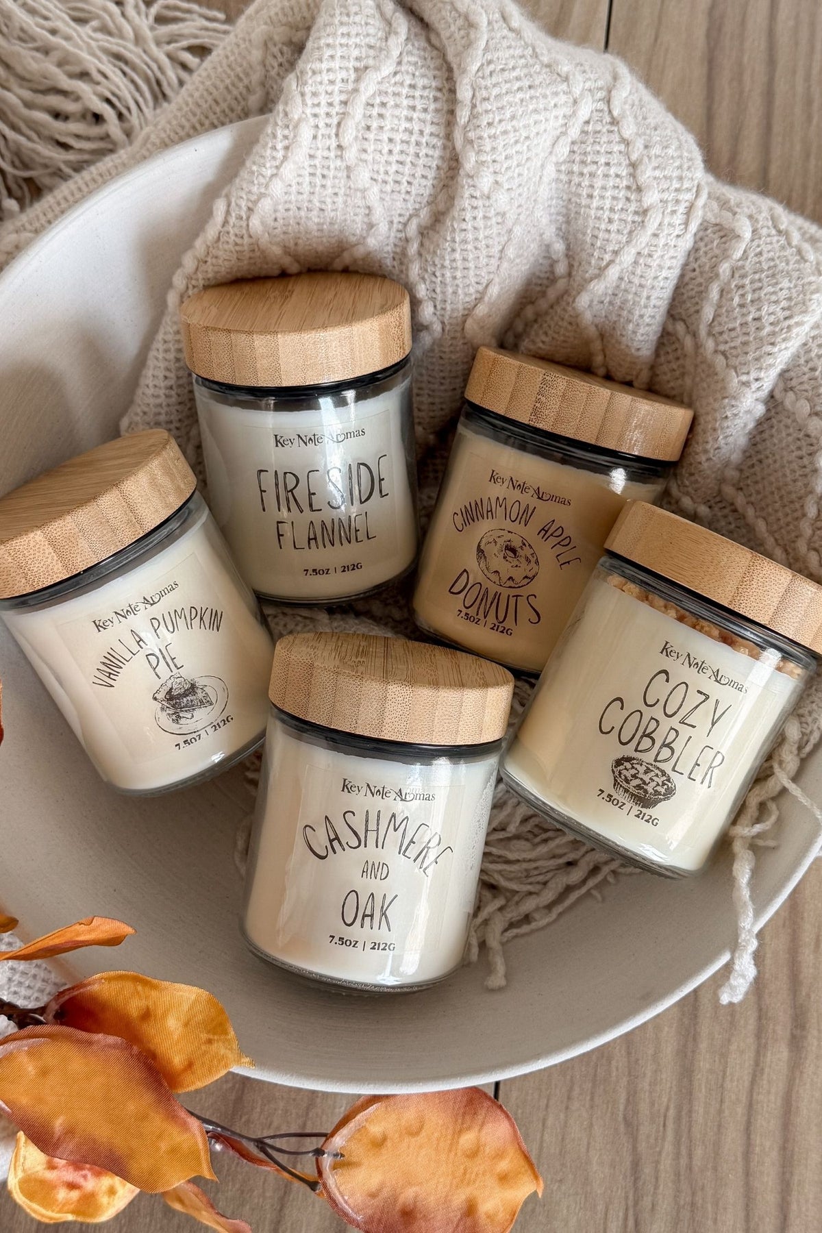 Five glass jar candles with wooden lids are arranged in a white bowl with a cream cable-knit throw and fall leaves on a wood surface. The labels read Vanilla Pumpkin Pie, Fireside Flannel, Cashmere and Oak, Cinnamon Apple Dumplings, and Cozy Cobblers.