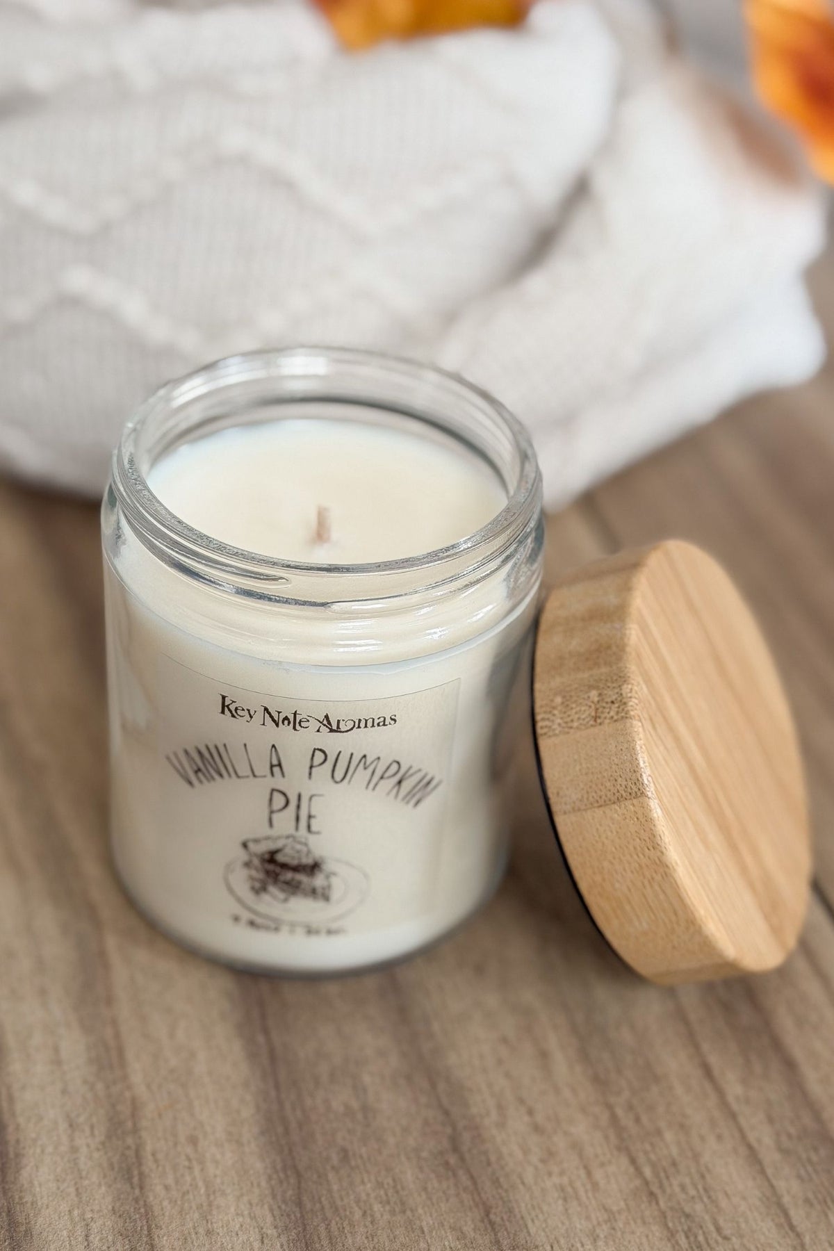 A small glass jar candle labeled 'Key Note Aromas Vanilla Pumpkin Pie' with white wax, shown next to a light wood lid on a wooden surface with a soft white textile in the background.