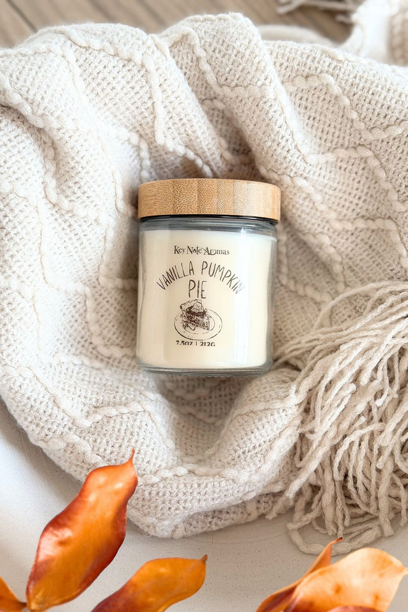 A jar candle labeled 'Vanilla Pumpkin Pie' by Key Note Aromas, styled on a cream cable-knit blanket with autumn leaves at the bottom.