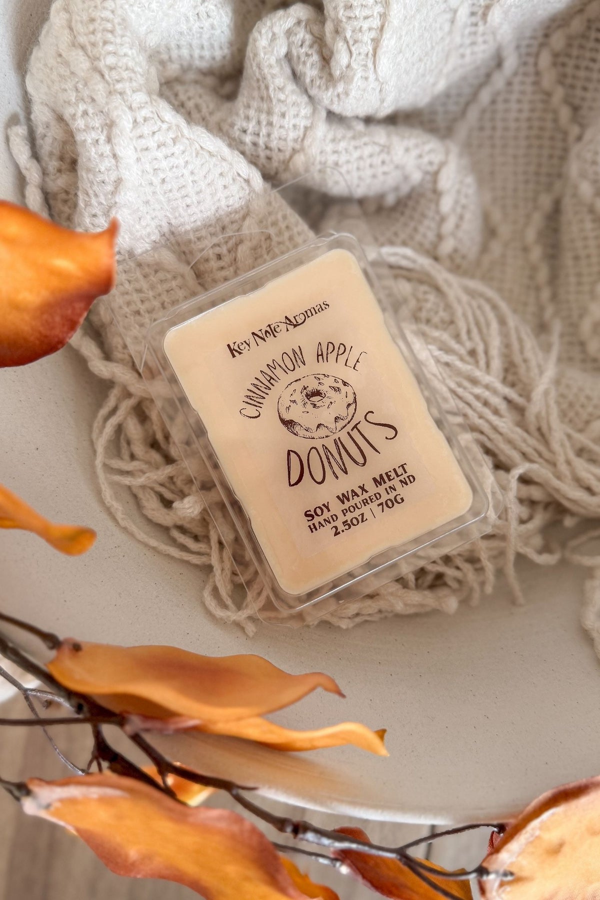 A square wax melt package labeled 'Cinnamon Apple Donuts' by Key note aromas, placed on a cream knit blanket with fall  leaves around it.