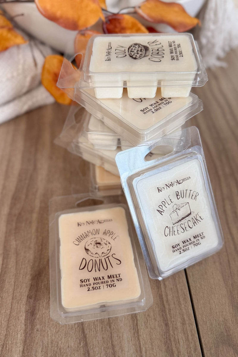 Three small packaged soy wax melts on a wooden surface. The products are in clear plastic clamshell packs with cream-colored labels and white wax inserts.