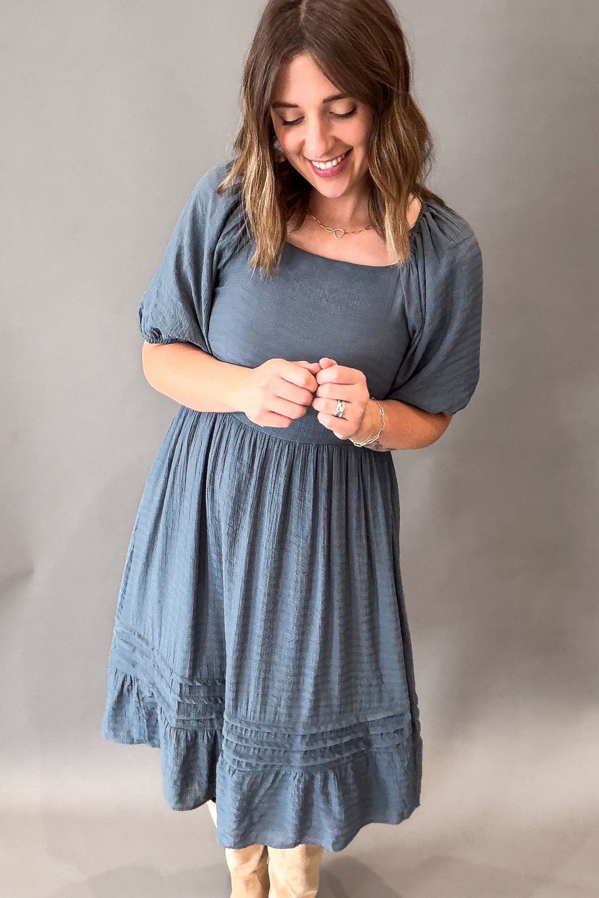 A woman wearing a dusty slate blue midi dress with short puff sleeves, a square neckline, and a gathered skirt, standing against a neutral gray studio background.