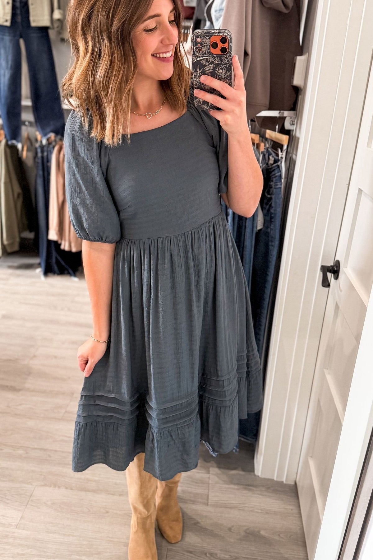 A woman takes a mirror selfie in a boutique wearing a dark gray short-sleeve midi dress with a tiered skirt and tan boots.