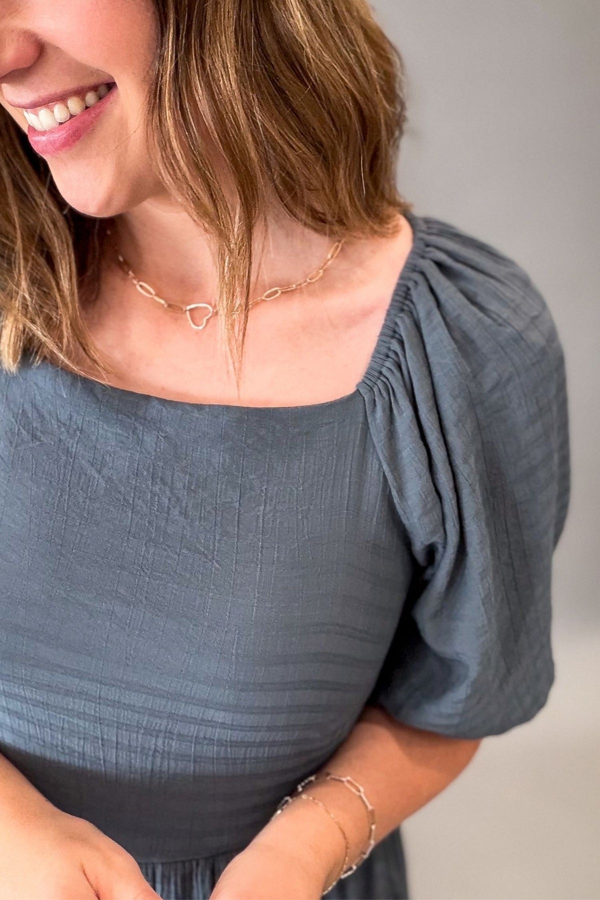 A close-up lifestyle shot of a woman wearing a slate blue-gray softly textured dress with a square neckline and voluminous puff sleeves. She is also wearing a delicate gold chain necklace with a heart pendant and a silver bracelet.