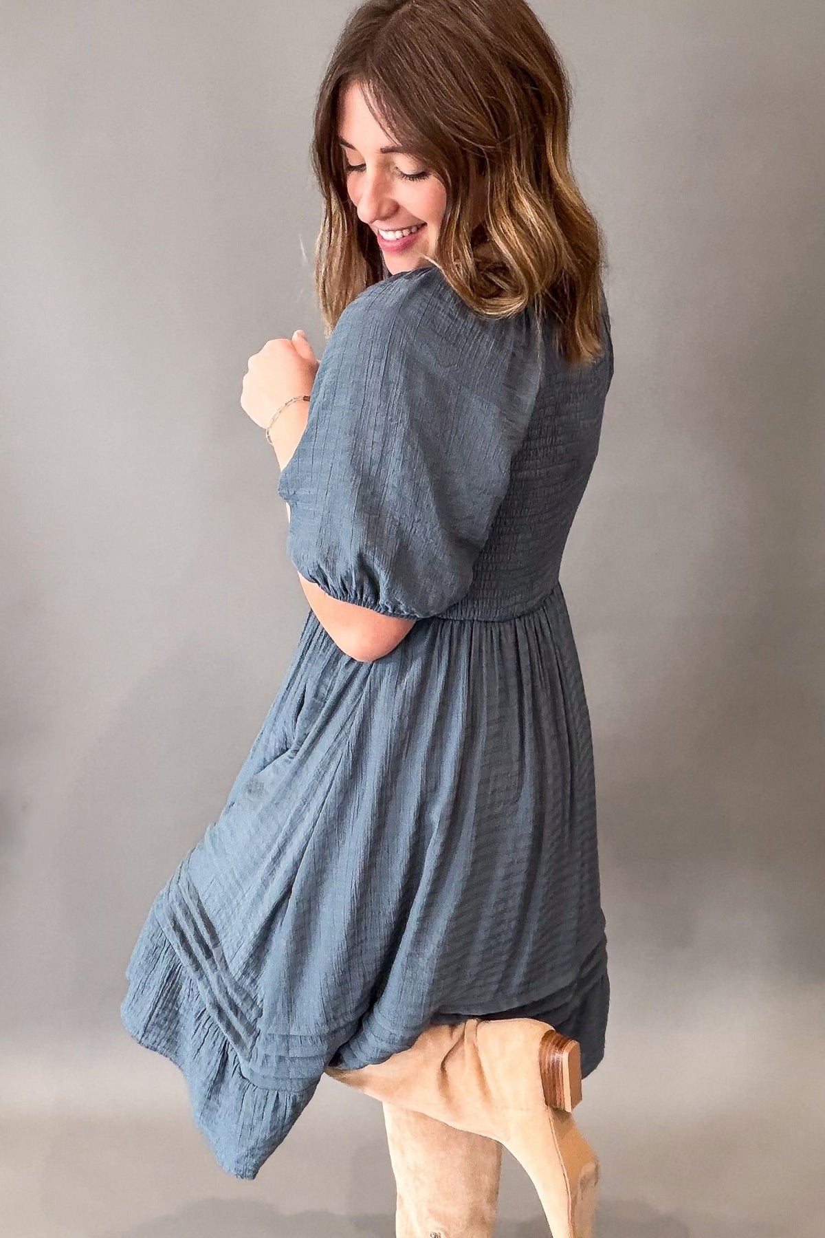 A woman wearing a dusty slate blue textured dress, photographed from the back and side against a neutral gray studio background.